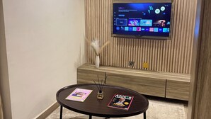 A modern living room with a flat-screen TV mounted on a wooden wall, a small table with magazines, and a comfortable sofa. - SAAFI Boutique Hotel (Abuja)