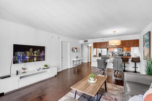A modern living room with a flat-screen TV, a dining area with a wooden table, and a kitchen with wooden cabinets.