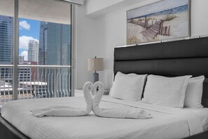 A hotel room with a large bed, white bedding, a heart-shaped towel arrangement, a painting of a beach scene, and a view of city buildings through a large window.