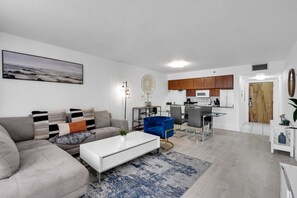 A modern living room with a grey sofa, a white coffee table, and a blue armchair. There is a kitchen area with wooden cabinets and a white microwave. - Elegant 1 BR Retreat in Brickell W/ Free Parking (Miami)