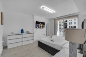 A modern bedroom with a bed, a dresser, a TV, and a window with a view of buildings.
