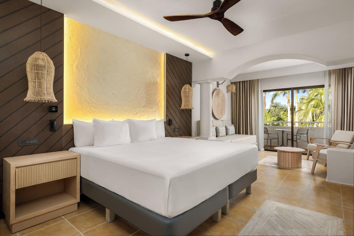 Una habitación de hotel moderna con una cama grande, ventilador de techo, zona de comedor con vistas al exterior y mesita de noche de madera.