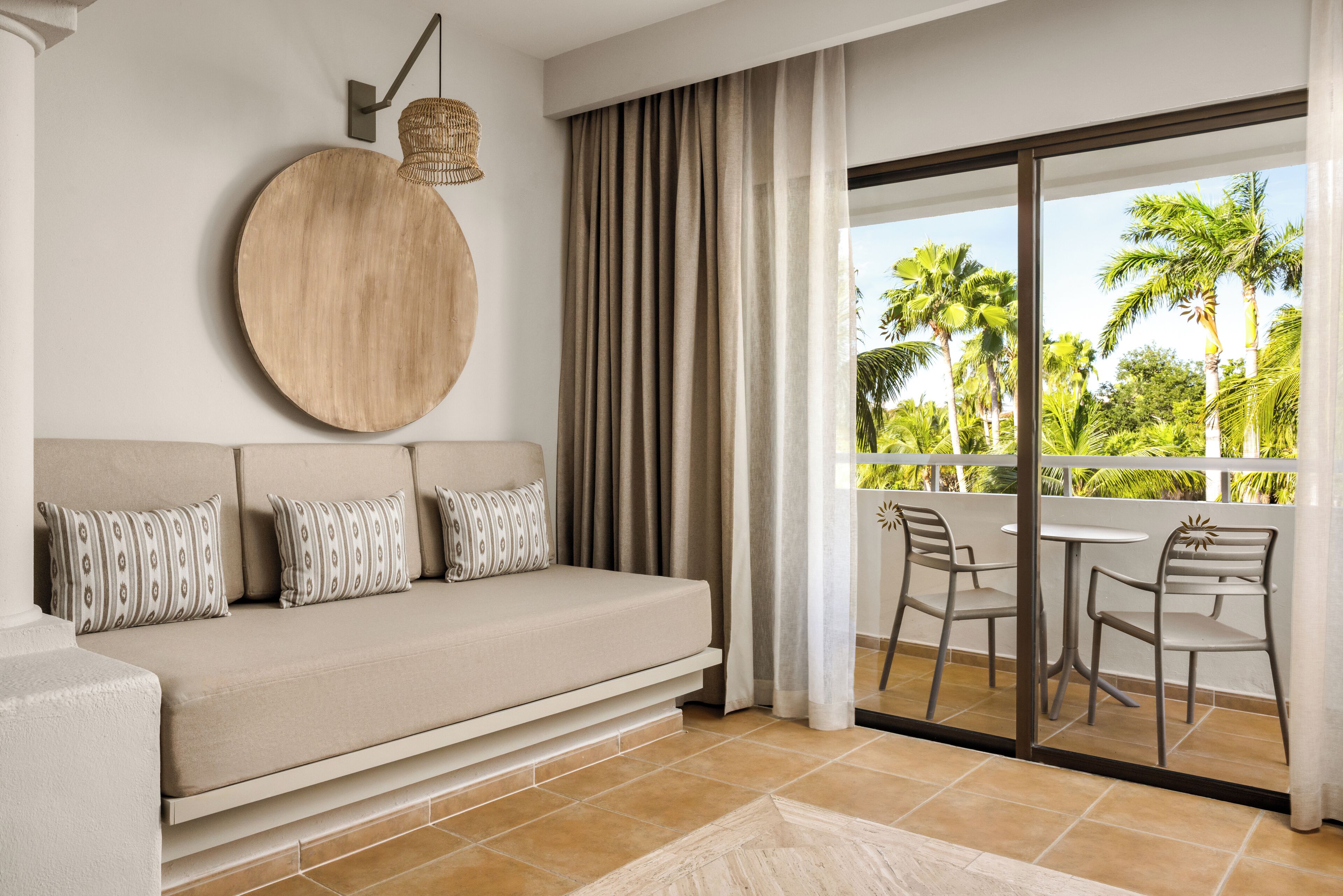 Una sala de estar moderna con un sofá, almohadones estampados, un adorno redondo en la pared y vistas a palmeras a través de un balcón.