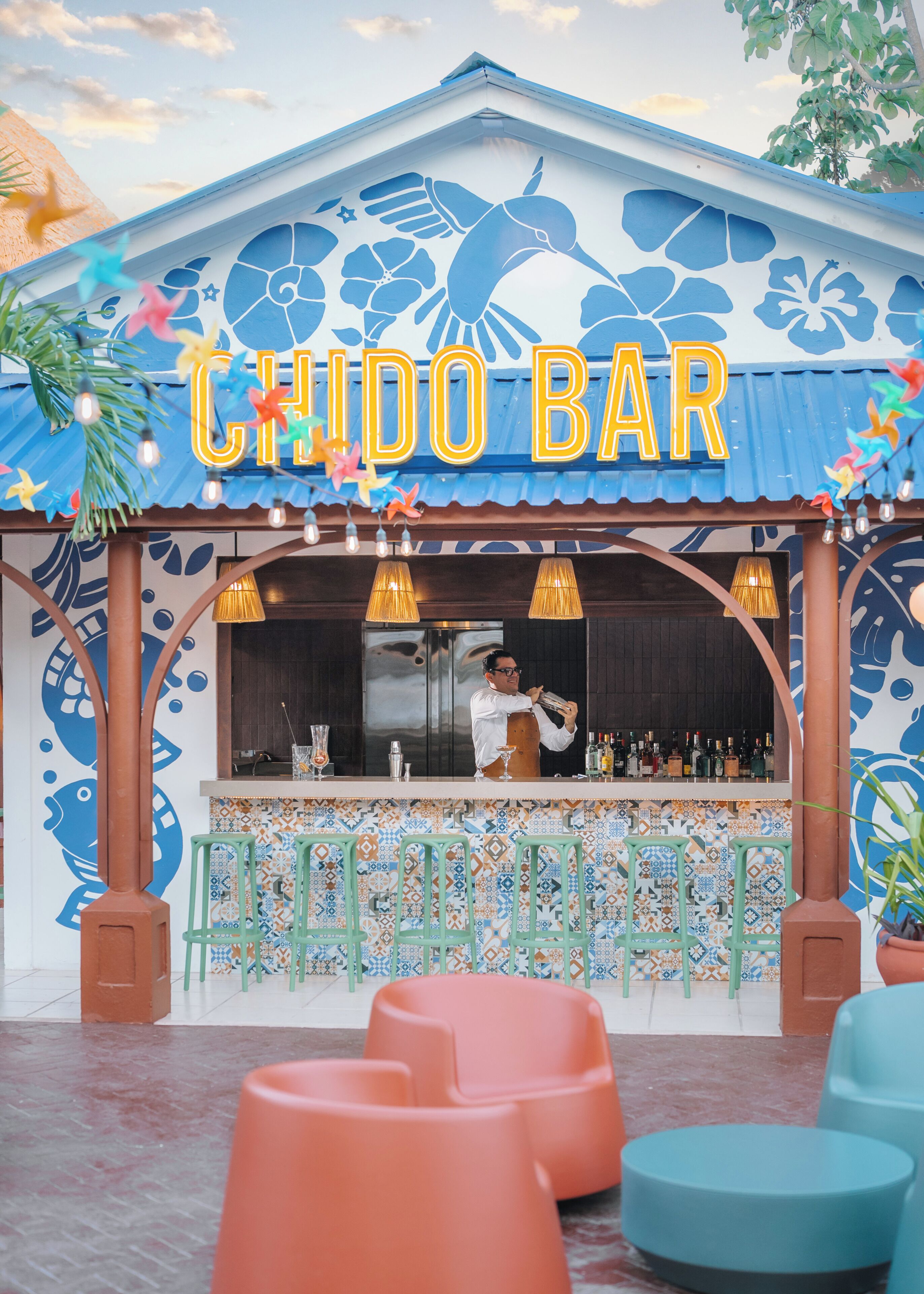 Un bar llamado 'CHIDO BAR' con un exterior colorido, un barman detrás de la barra y mesas al aire libre.