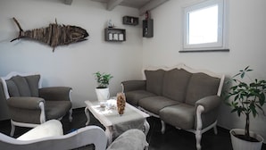 Olohuoneessa on harmaa sohva, kaksi nojatuolia, kasvilla koristeltu sohvapöytä ja seinälle asennettu kalaveistos.