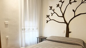 A neatly made bed with striped bedding and two folded towels, a small bedside table, and a wall-mounted tree decoration.