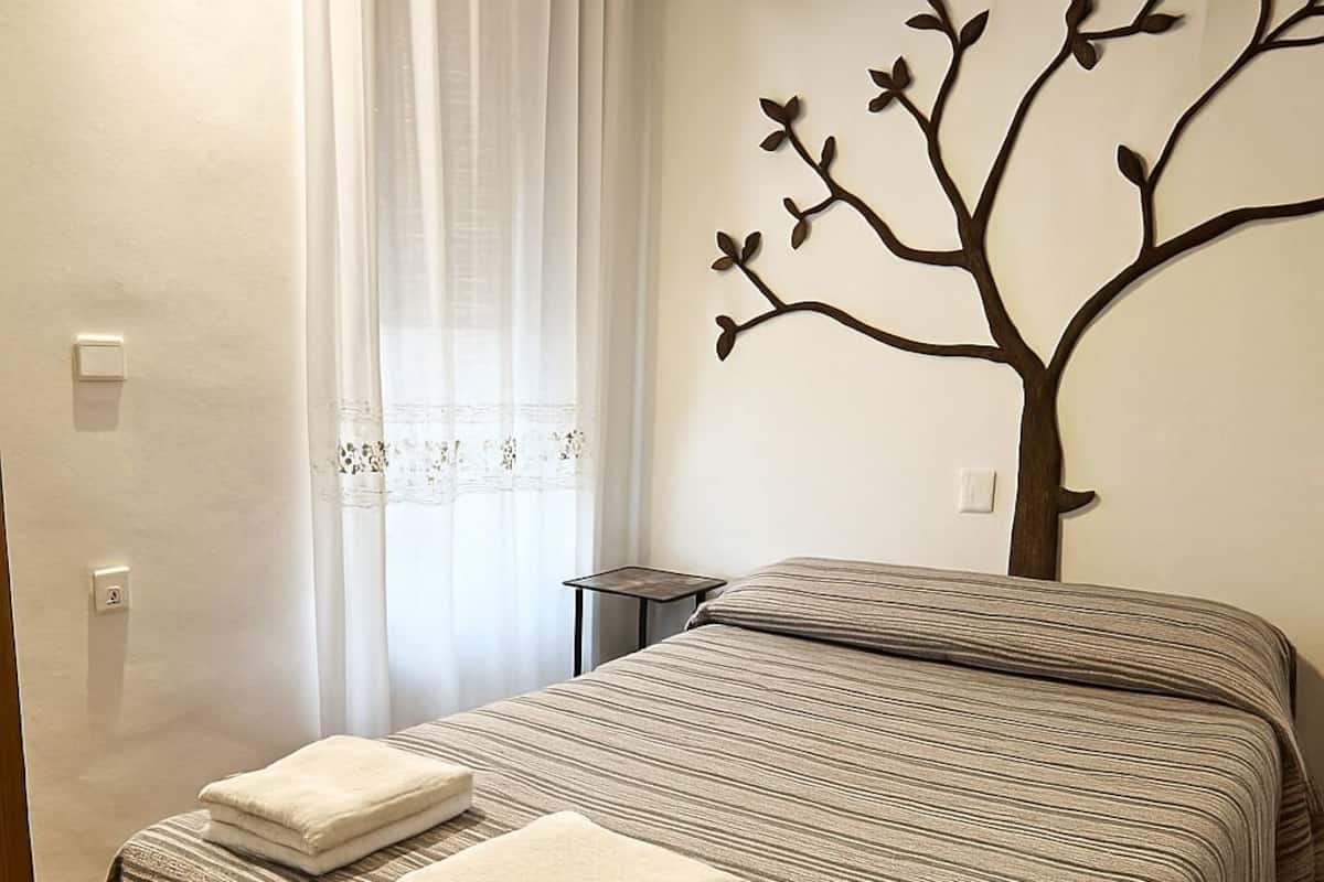 A neatly made bed with striped bedding and two folded towels, a small bedside table, and a wall-mounted tree decoration.