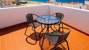 A rooftop terrace with a round table and four wicker chairs, overlooking a beach and the ocean.
