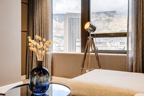 Habitación de hotel con cama, escritorio con un florero de flores secas, lámpara y ventana con vista a paisaje urbano y montañas.