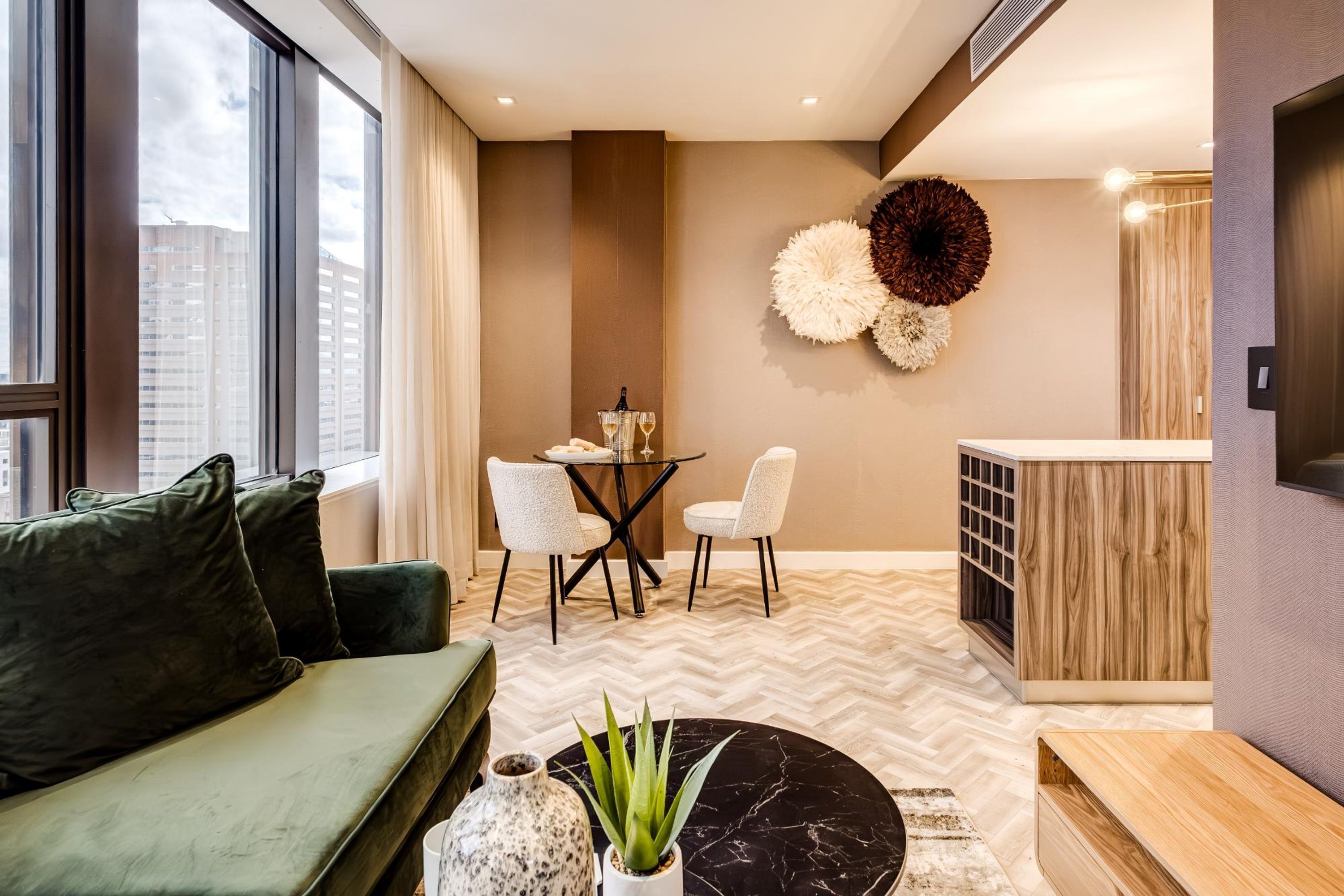 Una sala de estar moderna con un sofá verde, una mesa de centro redonda negra y un suelo con patrón de espiga. Se observa un comedor con una mesa redonda pequeña y sillas blancas. Se distingue una gran pieza decorativa en la pared.