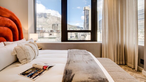 A hotel room with a large bed, a view of a cityscape through a window, and a red accent wall. - Trendy 1bed Modern Apt Cape Town City Suite 1601 (Cape Town)