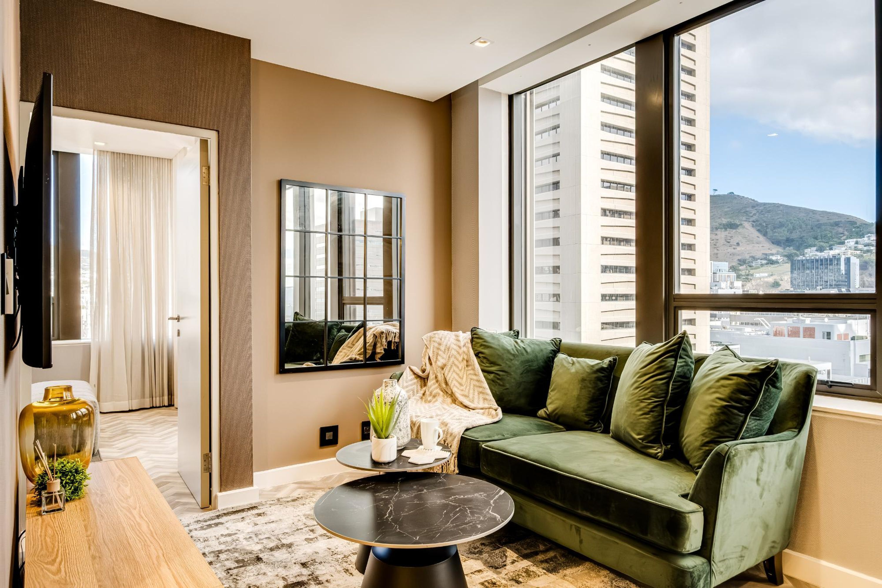 A modern living room with a green sofa, a round black table, and a large window offering a city view.