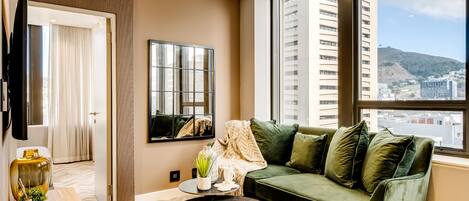 A modern living room with a green sofa, a round black table, and a large window offering a city view.