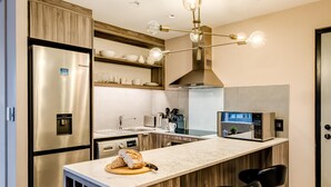 A modern kitchen with a stainless steel refrigerator, a built-in oven, a microwave, and a coffee machine. There is a wooden countertop with a cutting board and a loaf of bread. Two black bar stools are placed at the counter. - 1bed Apt in Cape Town CBD City Suite 1602 (Cape Town)