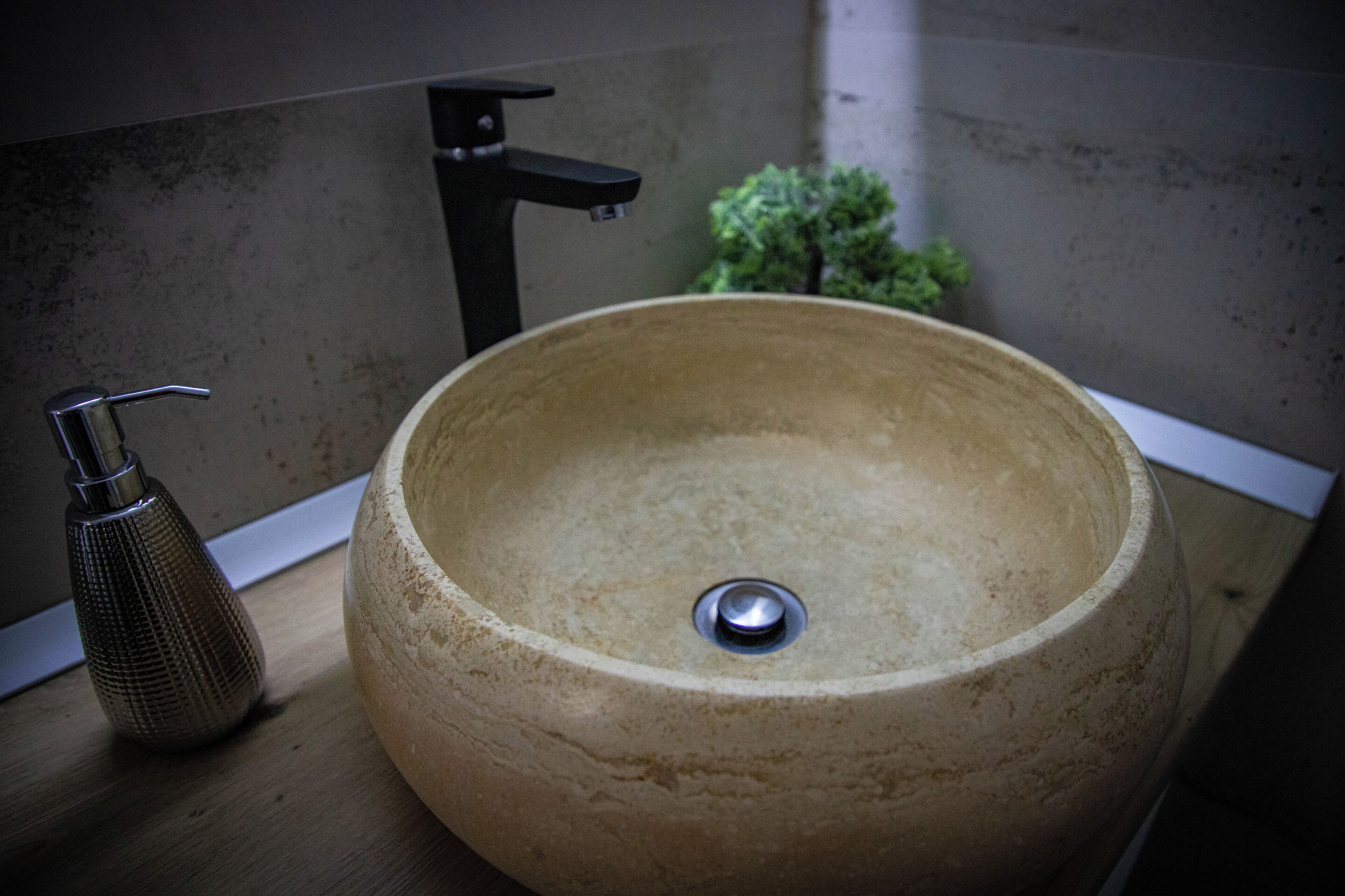 A large, round, beige ceramic sink with a modern faucet and a small, textured bottle on a wooden countertop.