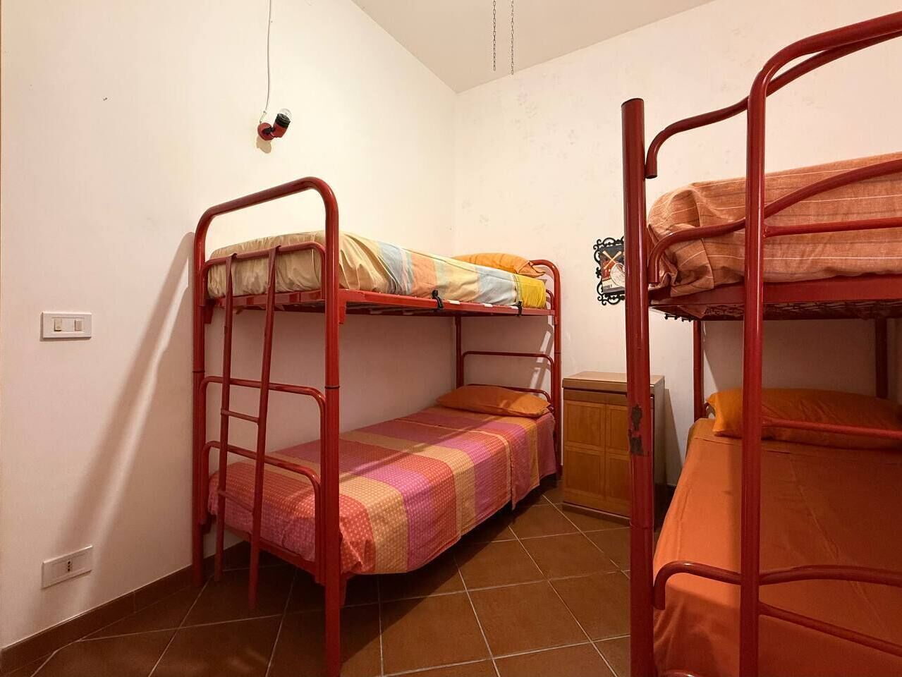 Een slaapzaal met stapelbedden, een klein houten kastje en een lichtschakelaar aan de muur.