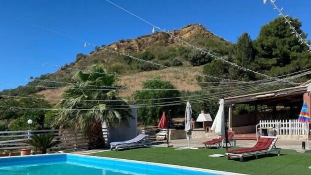 Una piscina con agua azul cristalina, rodeada por un patio con sillones de descanso y sombrillas. Al fondo, se divisa una colina con árboles y una cerca blanca de piquetes.