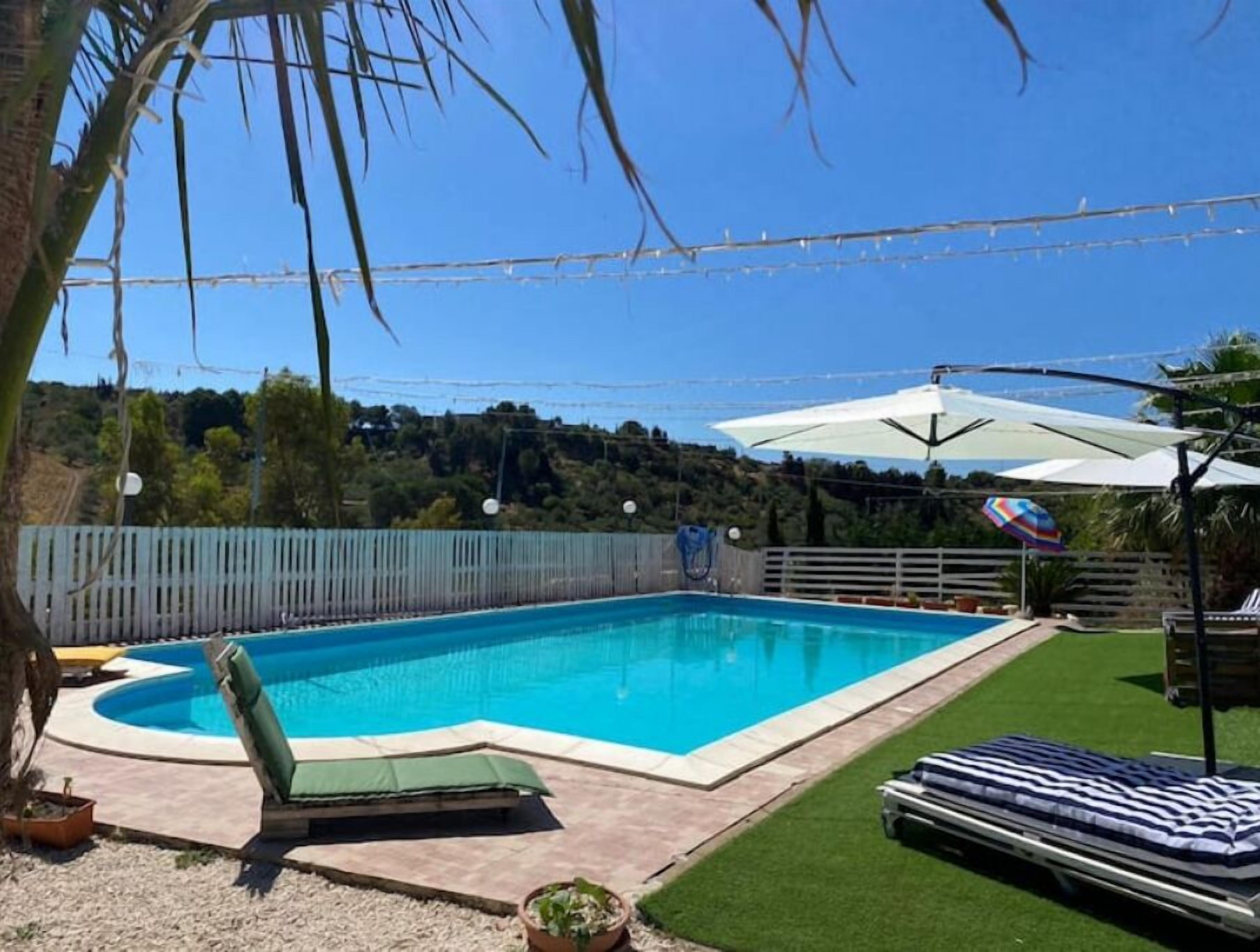 Une piscine rectangulaire bordée d’une pelouse verdoyante, le tout entouré d’une clôture en bois et agrémenté d’un parasol blanc.