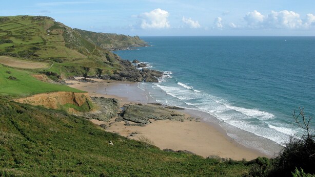 A coastal landscape with a beach, rocky shoreline, and green hills.
