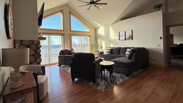 A spacious living room with a large gray sectional sofa, a small round coffee table, and a wooden floor. There are two windows with a view of a deck and a ceiling fan.
