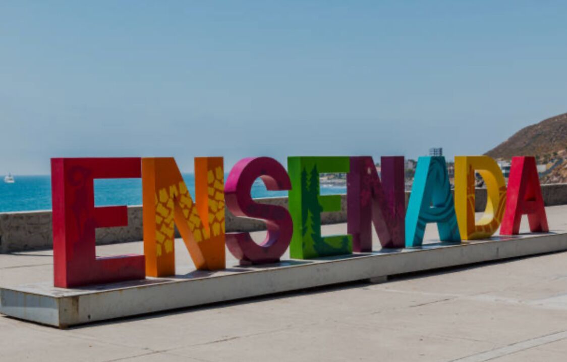 Large colorful letters spelling "ENSEÑADA" on a platform with a coastal background.