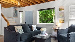 Ein modernes Wohnzimmer mit einem dunkelgrauen Sofa, einem passenden Sessel und einem Couchtisch. Ein Fenster bietet Blick auf einen Wald, eine Treppe und eine weiße Wand mit gerahmten Bildern.