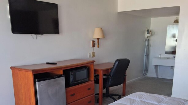 A hotel room with a bed, a desk with a microwave, a chair, a TV, a lamp, and a bathroom with a mirror and sink. - Coratel Inn & Suites by Jasper Anthony- 1 King Bed Non-Smoking (Anthony)