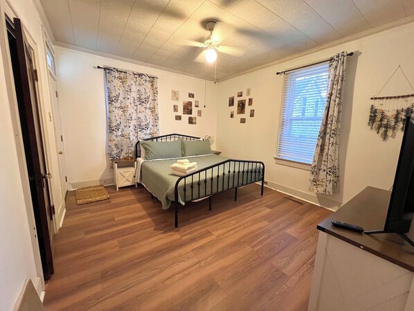 Un dormitorio con cama, ventilador de techo, ventana con cortinas y televisor instalado en la pared.