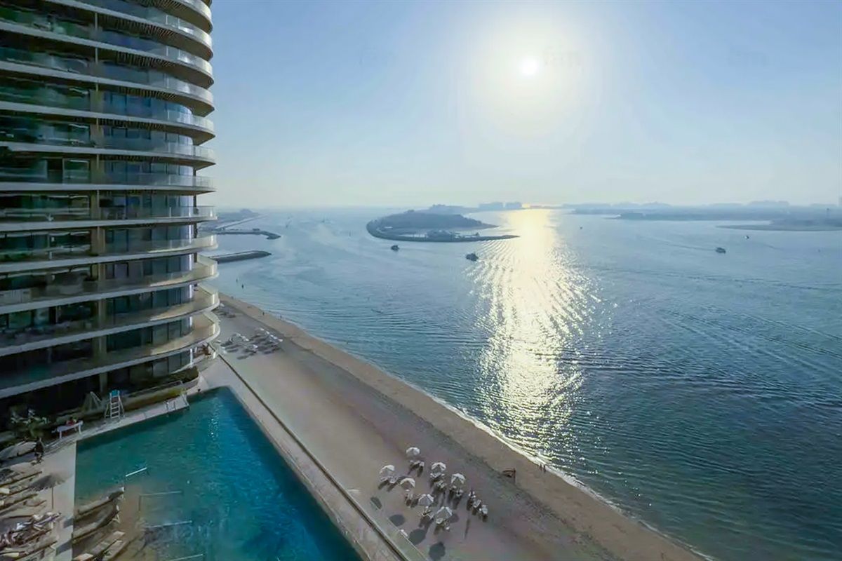 A high-rise hotel with a pool and beach area, overlooking a calm body of water with boats.