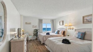 Hotellihuone, jossa on kaksi sänkyä, työpöytä, peili, merinäköala ja ranta-aiheinen sisustus.