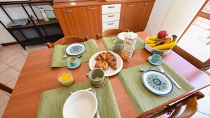 A dining table set with plates, bowls, and a fruit bowl, with a kitchen in the background. - Casa Vuvi - Two-room apartment very close to the beach. (Toscolano Maderno)