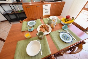 A dining table set with plates, bowls, and a fruit bowl, with a kitchen in the background.