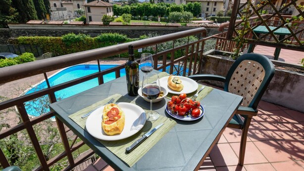 A table with food and drinks, a pool, and a view of buildings and greenery. - Casa Vuvi - Two-room apartment very close to the beach. (Toscolano Maderno)