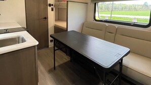 A compact RV interior with a kitchenette, a dining table, a sofa, and a window with a view of greenery. - NEW 2025 RV, 1 Bedroom WiFi AC in Serene Winterport with Coast and Bangor nearby (Winterport)