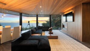 Ein modernes Wohnzimmer mit einer großen Eckcouch, einem Couchtisch und einem an der Wand montierten Fernseher. Ein großes Fenster bietet Blick auf den Ozean und einen Sonnenuntergang.