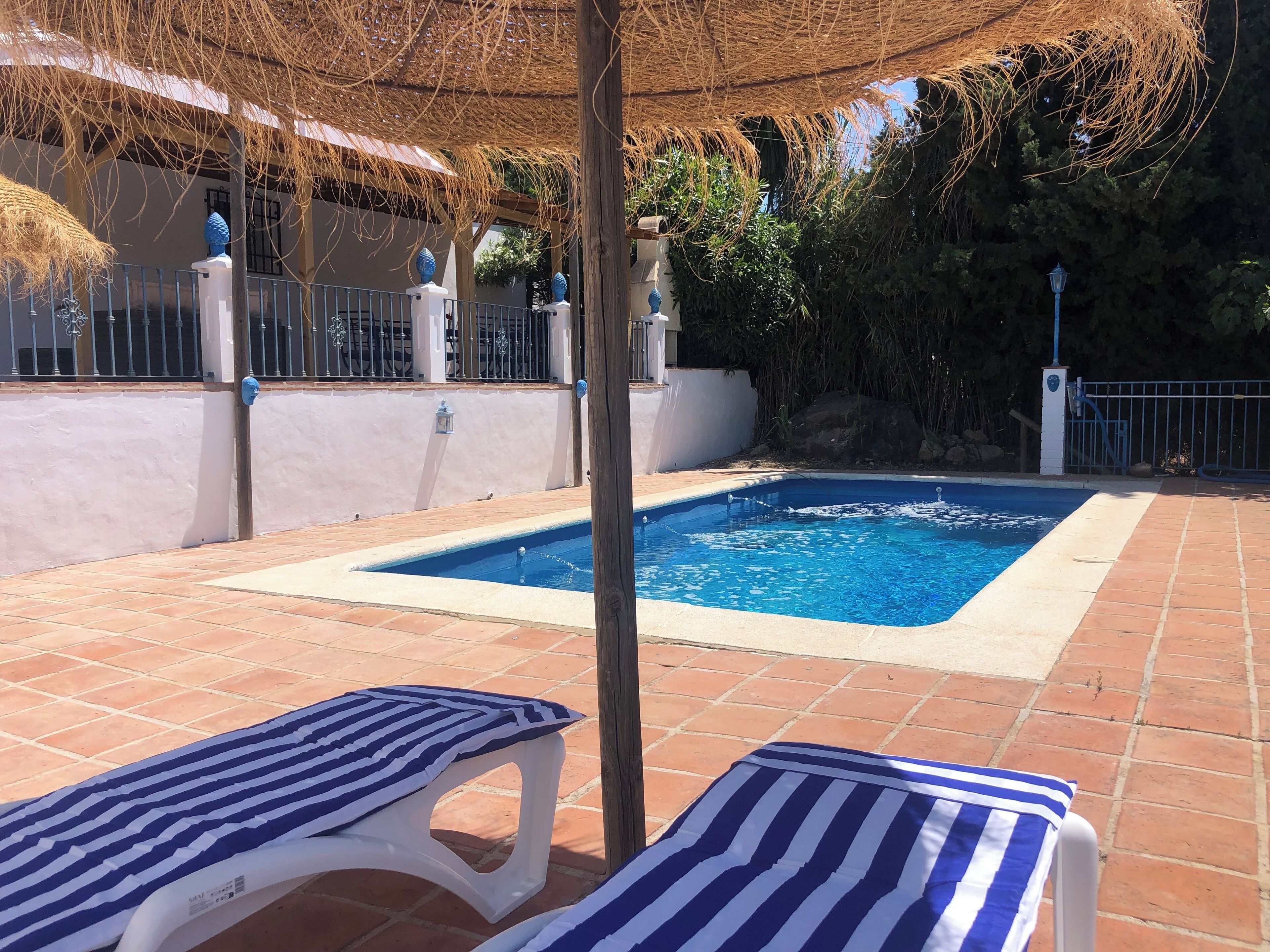 A swimming pool with a tiled deck, two striped lounge chairs, and a thatched roof structure.