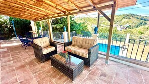 Eine überdachte Terrasse mit Korbmöbeln, einem Glastisch und Blick auf einen Swimmingpool und eine hügelige Landschaft.