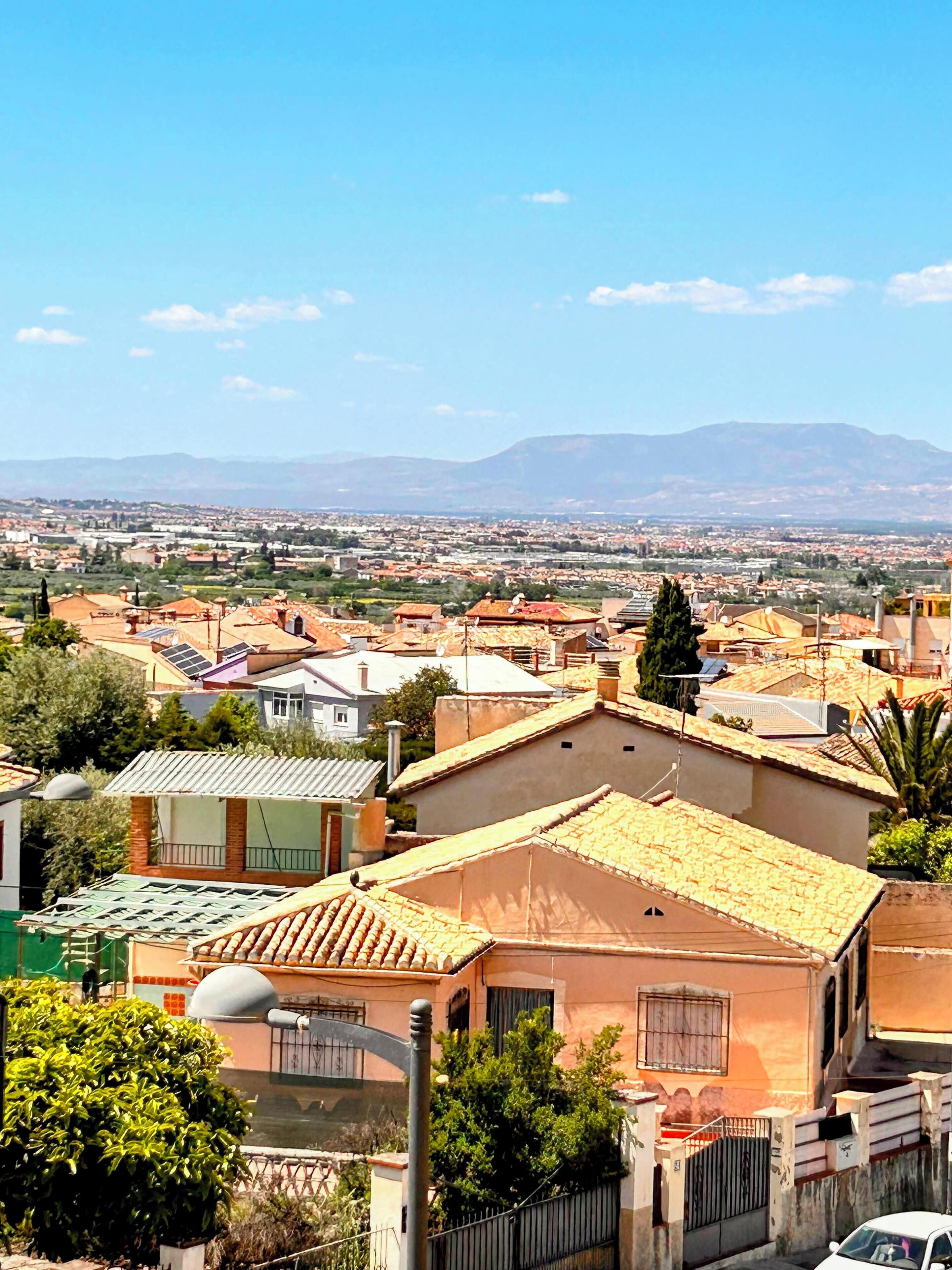A residential area with terracotta-roofed houses, a clear sky, and distant mountains.