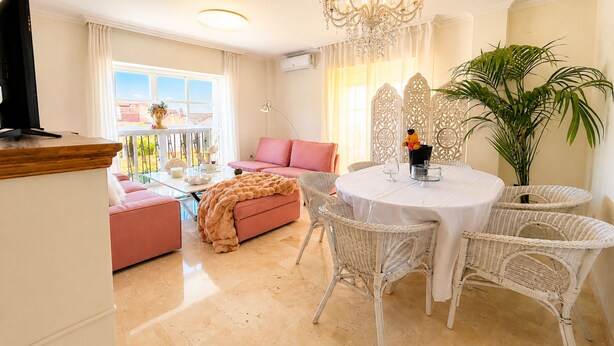 Una sala de estar con un sofá rosa, una mesa redonda de comedor con sillas de mimbre blanco, una araña y una planta grande en maceta.