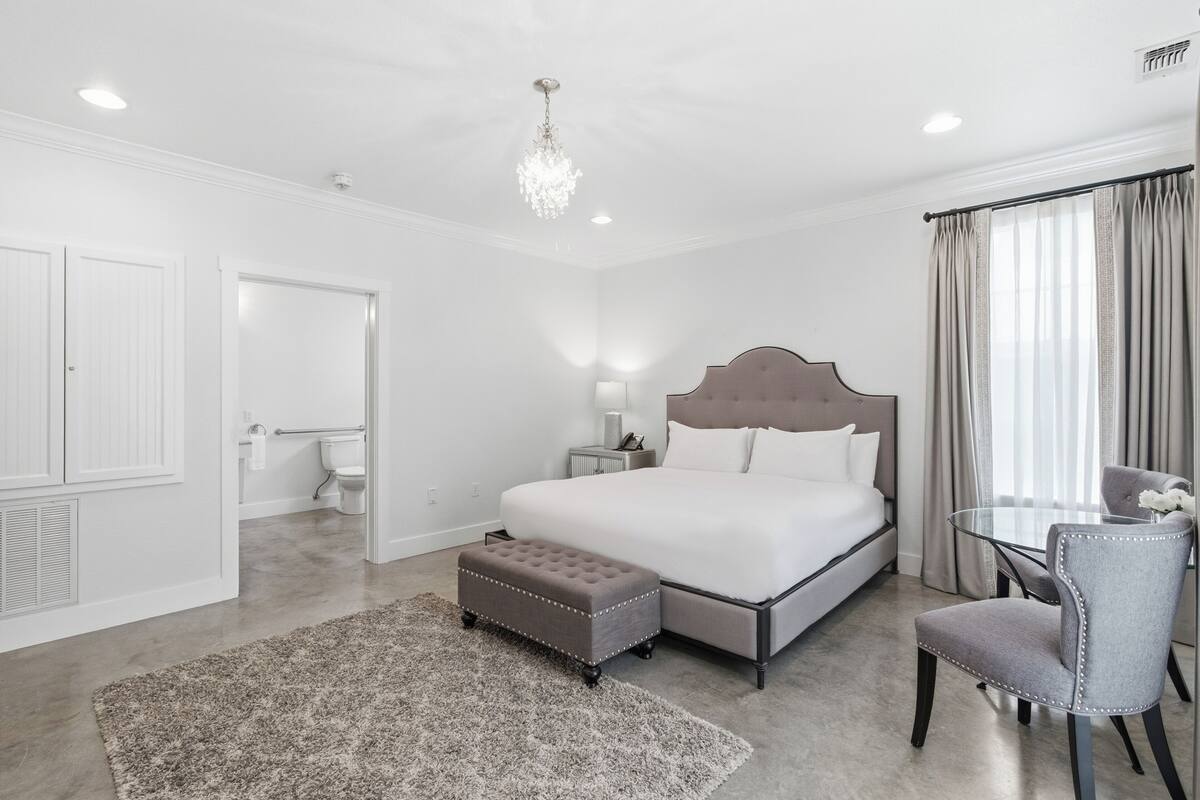 A bedroom with a bed, a bedside table, a chair, a small table, a chandelier, and a window with curtains.