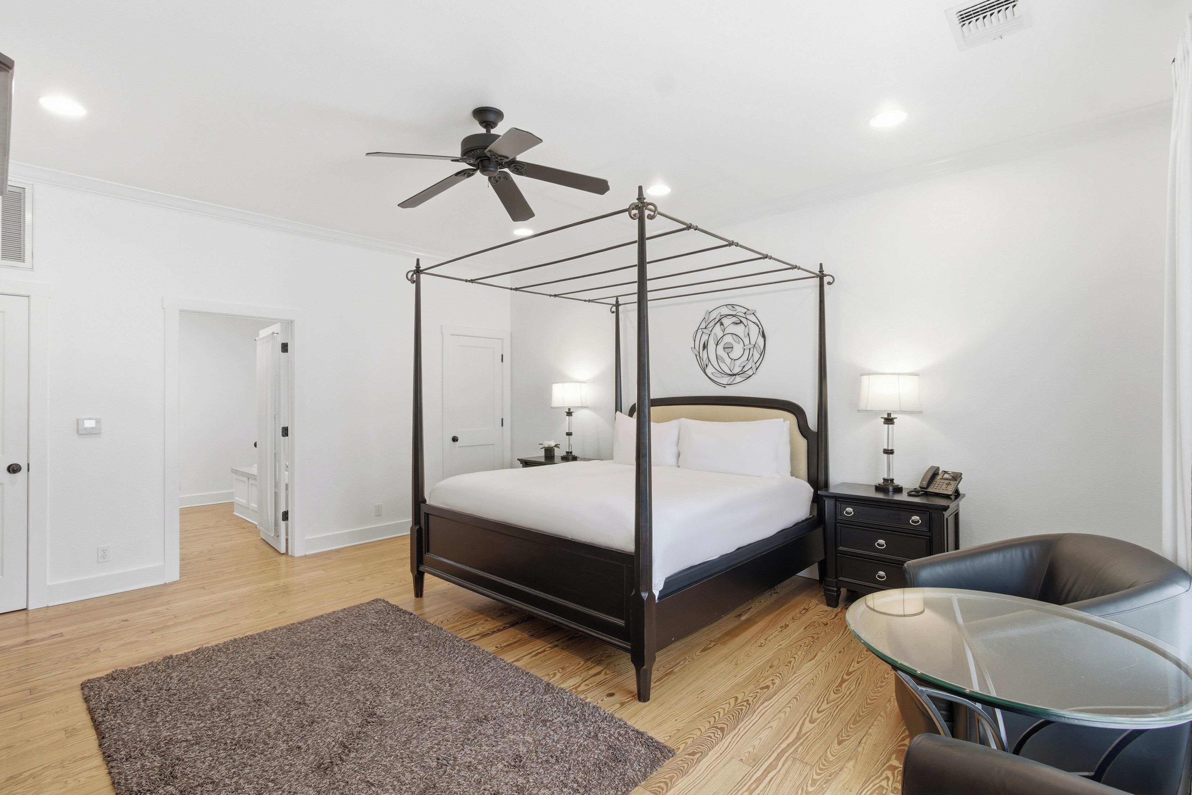 A bedroom with a four-poster bed, a ceiling fan, bedside lamps, a glass table, and a chair.
