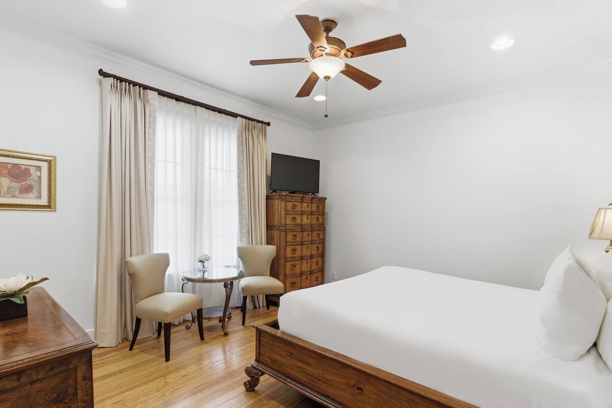 A hotel room with a bed, a ceiling fan, a TV, a wooden dresser, a desk, and a chair.