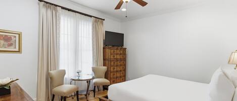 A hotel room with a bed, a ceiling fan, a TV, a wooden dresser, a desk, and a chair.