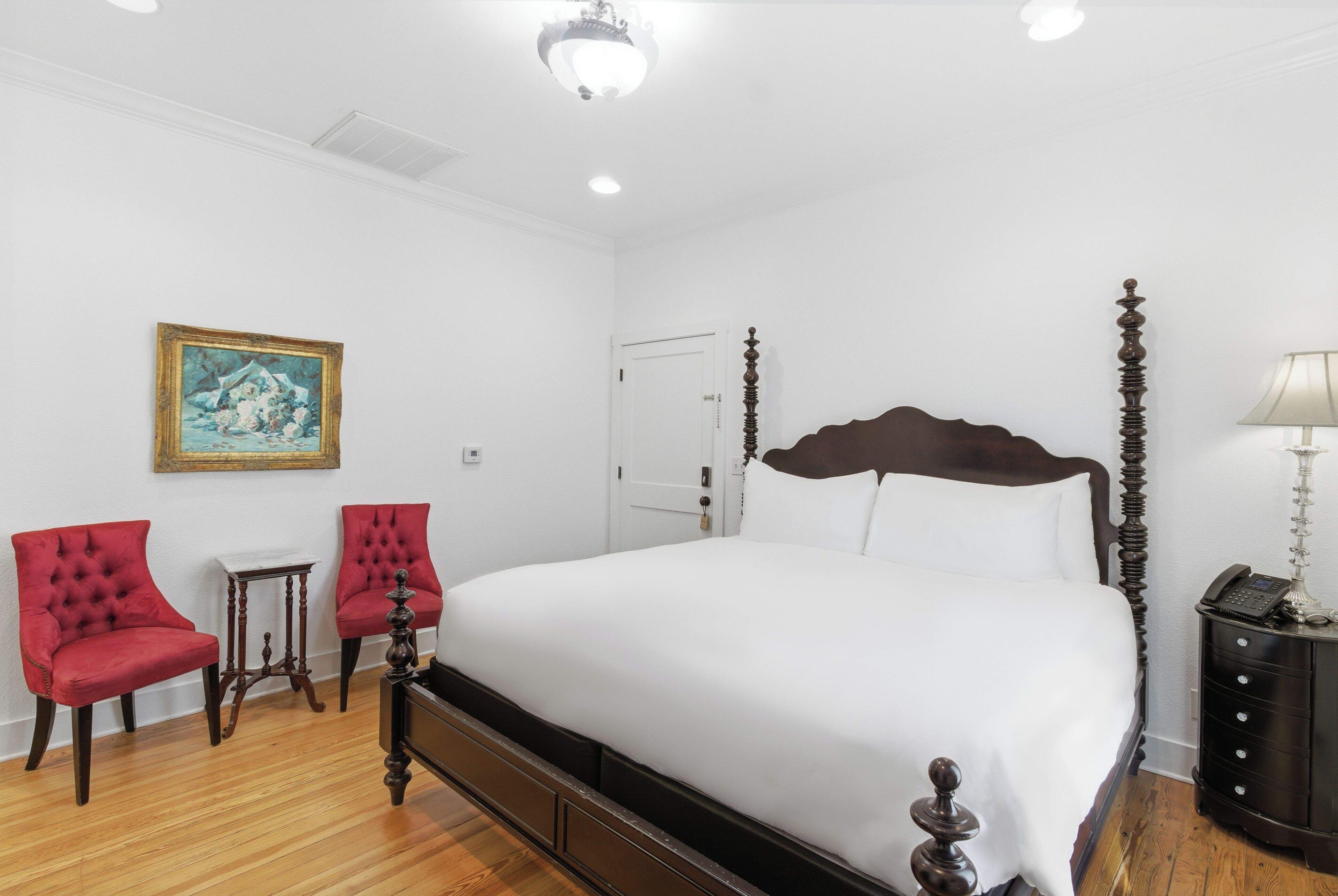 A bedroom with a large bed, two red chairs, a small table, a painting on the wall, and a nightstand with a lamp.