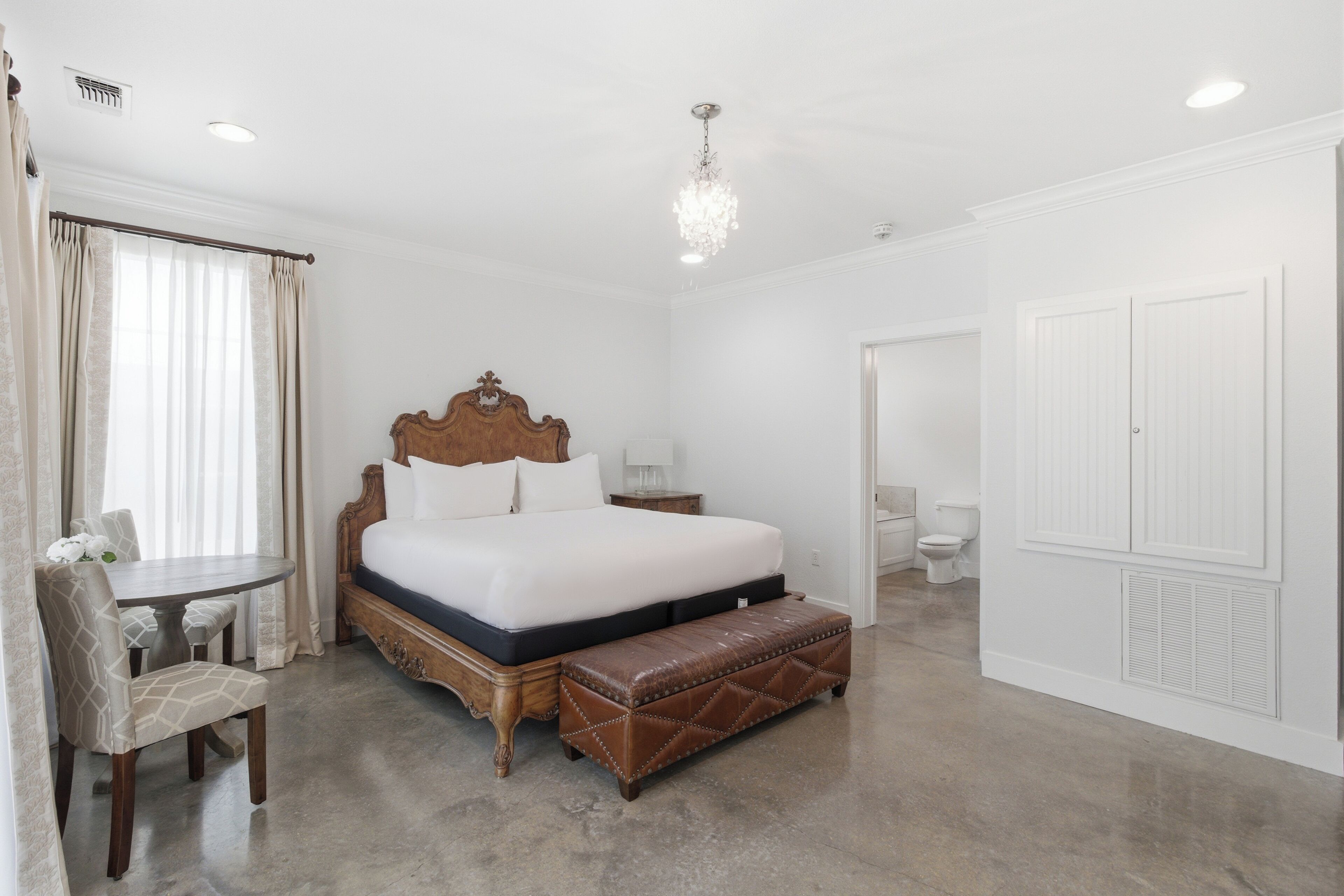 A bedroom with a large bed, a wooden headboard, a bench, a chair, a bedside table, and a chandelier.