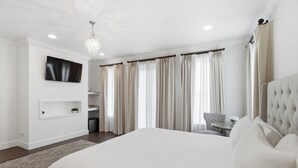 A modern bedroom with a large bed, a TV mounted on the wall, a chandelier, and a dining area with a chair and table. - Emigrant's Inn (Fredericksburg)