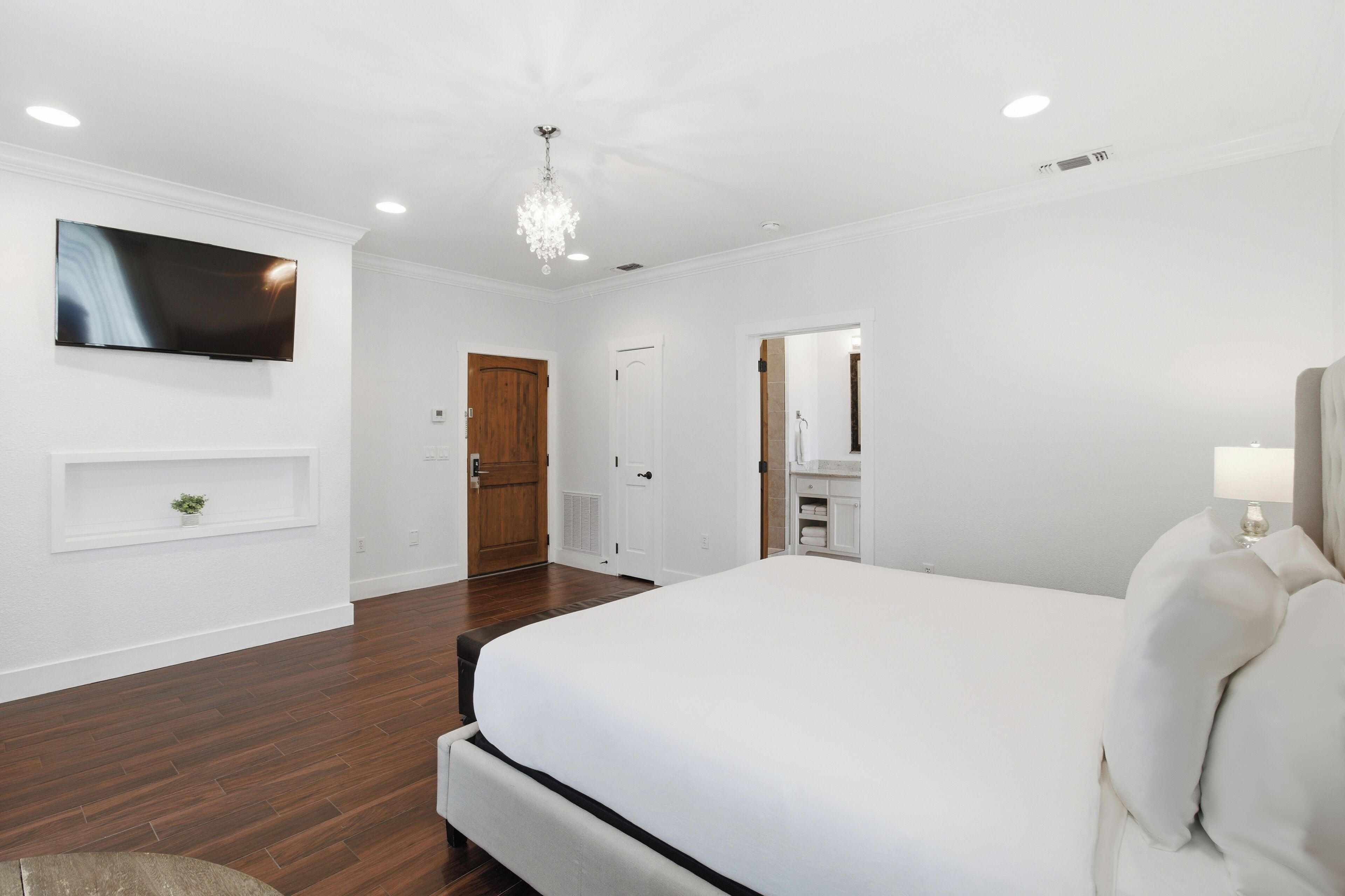 Un dormitorio con una cama grande, un televisor montado en la pared, una puerta de madera y una planta pequeña en un estante.
