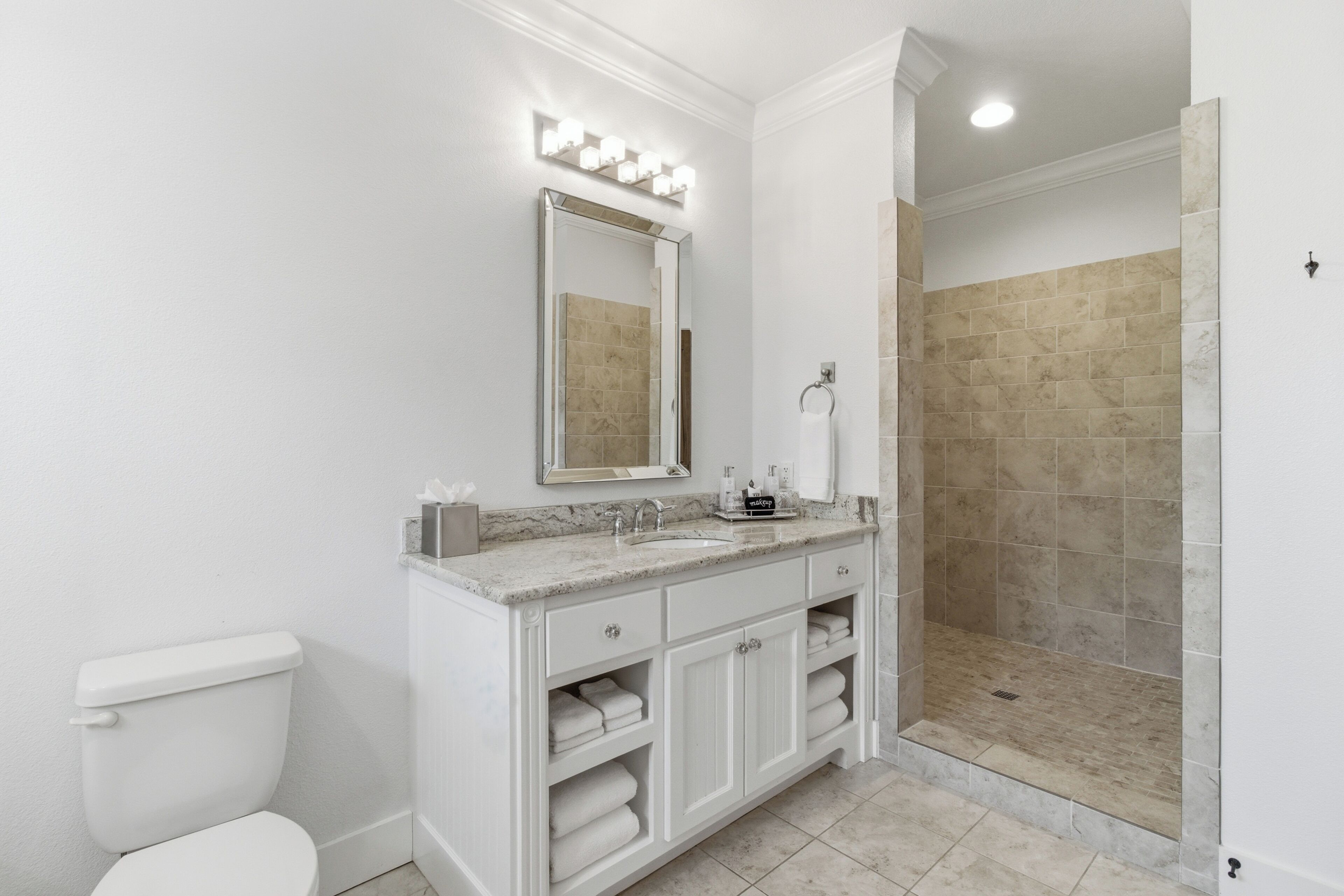 A bathroom with a toilet, a sink, a mirror with lights, a shower area, and a vanity with cabinets.