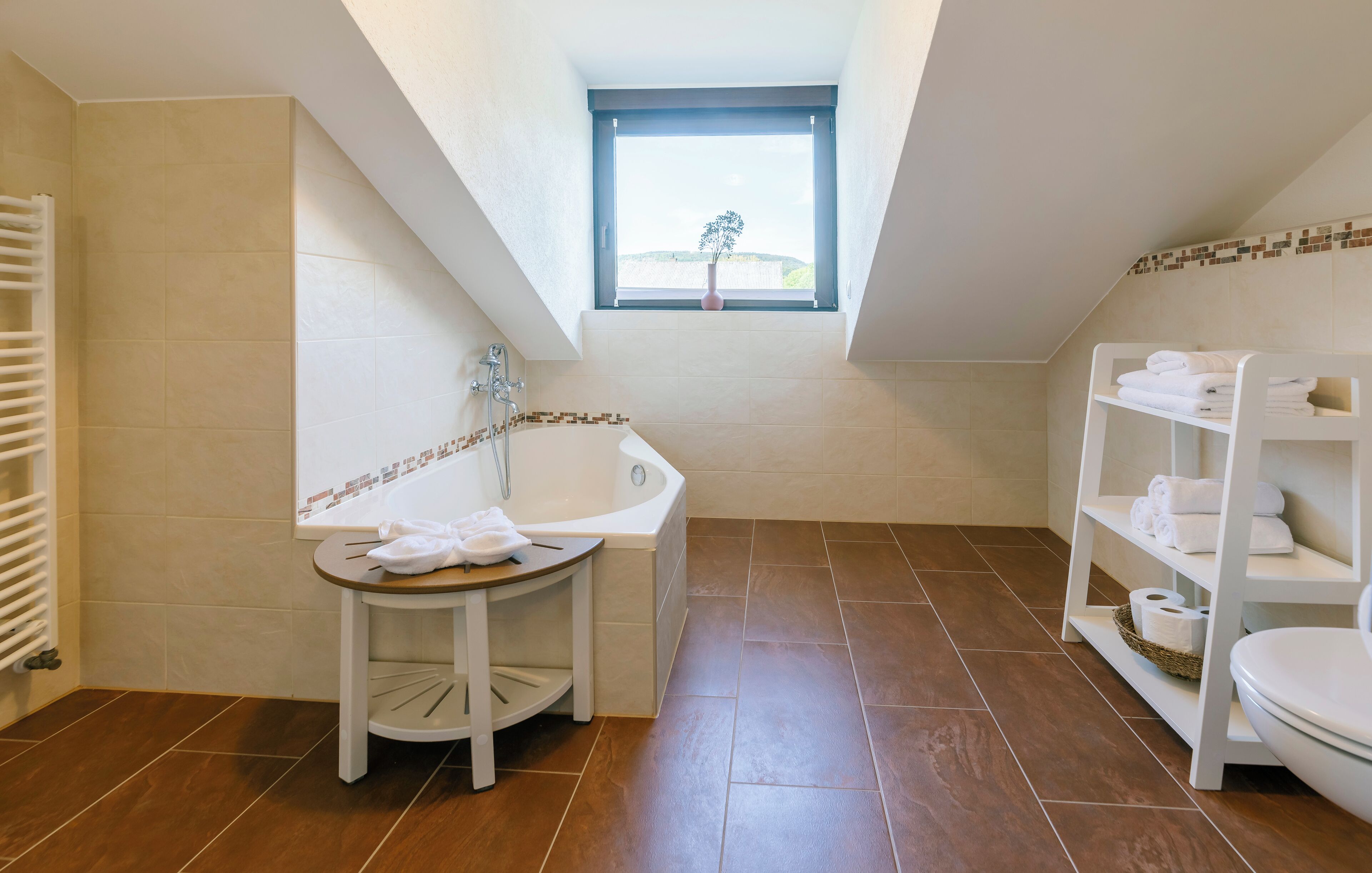 Un baño moderno con una bañera independiente, un tocador pequeño con un taburete, un estante con toallas y un ventanal con vistas a la vegetación.