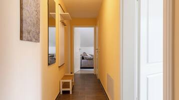 A narrow hallway with a bench, a door at the end, and a room with a bed and a window.